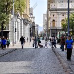 Pessoas caminham por uma rua movimentada de Santiago, no Chile, cercada por edifícios históricos.